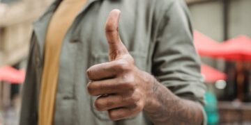 Crop faceless young black male in casual outfit standing on city street and showing thumb up gesture