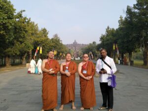 This image depicts a group of Buddhist aspiring monks and a man in traditional clothing standing in front of the Borobudur temple complex in Indonesia. The temple can be seen in the background, surrounded by lush greenery. The monks are wearing orange robes and appear to be part of a religious or cultural event or gathering at the site. The man in the foreground is also dressed in traditional attire, he is Ambassador from Sri Lanka who was present at the celebration of the Indonesia Tipitaka Chanting 2024 event The scene captures a moment of cultural and spiritual interaction at the renowned Borobudur temple, which is a significant Buddhist archaeological site and UNESCO World Heritage location.