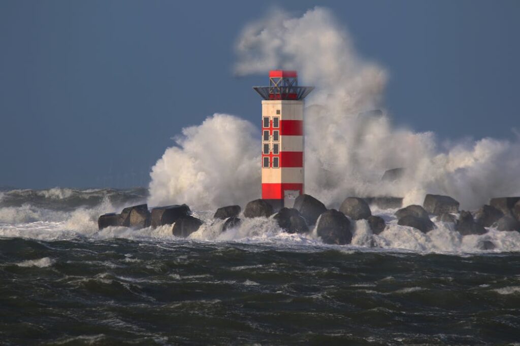 Red and white lighthouse with waves crashing against rocks under a stormy sky.