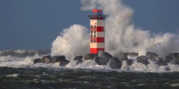 Red and white lighthouse with waves crashing against rocks under a stormy sky.