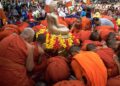 Monks in orange robes conduct a religious ceremony around a golden Buddha statue outdoors.