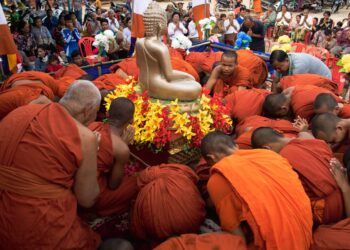 Monks in orange robes conduct a religious ceremony around a golden Buddha statue outdoors.