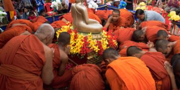 Monks in orange robes conduct a religious ceremony around a golden Buddha statue outdoors.